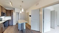 This well-appointed single-floor residence showcases a thoughtfully designed open-concept layout that maximizes functionality and flow. The kitchen serves as a central highlight, featuring a granite-topped island with sleek metal legs, providing both additional workspace and a casual dining or gathering spot. Elegant pendant lighting with cone-shaped fixtures hangs above the island, adding a modern touch to the space. The kitchen connects seamlessly to the rest of the unit, allowing for easy movement throughout. Hardwood-style flooring runs throughout the main living areas, lending warmth and a cohesive aesthetic. The layout includes multiple rooms accessible from a central hallway, each framed by clean white panel doors that contribute to the crisp, contemporary feel. Closet space is thoughtfully integrated, with at least one visible walk-in or reach-in closet offering practical storage solutions. The walls are finished in a neutral, light tone that enhances the brightness of the space. Ceiling vents suggest a central HVAC system for year-round comfort. Overall, the residence presents a clean, modern aesthetic with practical design elements that cater to everyday living.