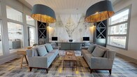 Located at 2155 Morris Basket Blvd in North Charleston, SC, this single-floor community lounge and business center is a standout example of thoughtful interior design. The space is anchored by a dramatic floor-to-ceiling nature-inspired mural depicting a stag, woodland creatures, and botanical illustrations in delicate line art, creating an artistic focal point that sets the tone for the entire room.

A built-in workstation finished in muted sage green runs along the mural wall, housing multiple computer terminals and providing a functional business center within the lounge. Flanking the workstation are open, X-pattern display shelving units that add architectural interest and depth. Overhead, oversized drum pendant lights with warm gold-toned interiors cast an inviting ambient glow throughout the space.

The lounge area features multiple seating arrangements, including gray upholstered sofas, accent chairs with patterned fabric, and a rustic wood trestle table, all set atop a bold plaid-patterned area rug in shades of gray, yellow, and navy. Large windows along one wall flood the space with natural light and offer views of the surrounding exterior. The entryway corridor is accented with board-and-batten wall paneling in soft gray and framed dragonfly artwork, while a sliding barn door adds a charming transitional element leading into adjacent areas.