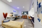 The Lounge at 6565 McCallum Blvd in Dallas, Texas is a thoughtfully designed single-floor amenity space that blends comfort, functionality, and modern style. The open layout is anchored by a bright, white-walled interior with light wood-look flooring throughout, creating a clean and welcoming atmosphere.

A skylight in the vaulted ceiling section floods the lounge area with natural light, complementing the recessed lighting overhead. A signature blue neon sign reading "The Lounge" serves as a focal point on the wall, adding a vibrant, contemporary touch. The seating area features a tufted cushioned sofa and a pair of leather lounge chairs arranged around a soft area rug, providing a relaxed environment for residents to gather.

A long wall-mounted desk with bar-height seating and workstation equipment offers a dedicated business center area, ideal for remote work or study. Adjacent to it, a decorative wood cabinet adds warmth and texture to the space.

The kitchen area is fully equipped with stainless steel appliances — including a refrigerator, electric range, and microwave — paired with white cabinetry and a tile backsplash. A navy blue island with a white quartz countertop and integrated sink is lined with leather bar stools, functioning as both a prep area and a casual gathering spot. French doors provide access to the exterior, further enhancing the open and connected feel of the space.