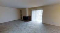 This single-floor space offers a clean, functional layout designed for comfortable everyday living. The living room serves as the central gathering area, featuring plush wall-to-wall carpeting that adds warmth underfoot. A corner fireplace with a tiled hearth provides a cozy focal point, complemented by built-in shelving along the adjacent wall — ideal for displaying books, décor, or media equipment. Large sliding glass doors dressed with vertical blinds flood the room with natural light and offer direct access to an outdoor area, creating a seamless connection between interior and exterior spaces. The neutral, light-toned walls throughout contribute to a bright and open ambiance, making the space feel airy and versatile. The overall design is straightforward and practical, with a palette that provides a blank canvas for personalization. The single-floor layout ensures easy accessibility and a cohesive flow throughout the home.