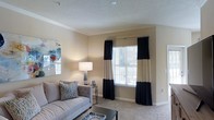 This single-floor space features a well-appointed living room designed with a clean, contemporary aesthetic. The room is finished with wall-to-wall carpeting in a neutral tone, complemented by smooth beige walls and white baseboards that create a warm, inviting atmosphere. Crown molding adds an elegant architectural detail along the ceiling line, which features a textured finish overhead.

A large window serves as the focal point of the room, dressed with bold navy and cream color-blocked curtains that frame the natural light filtering in through horizontal blinds. The window offers a glimpse of an outdoor balcony or deck area beyond. The living area is furnished with a tufted gray sofa accented by patterned throw pillows, paired with a sleek glass coffee table. A glass-topped side table holds a modern table lamp, providing warm ambient lighting. A piece of colorful abstract wall art adds a vibrant touch to the space.

Overall, the living room balances comfort and style, with thoughtful design details that create a functional yet visually appealing environment.