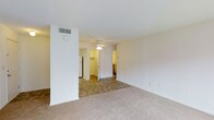 This single-floor residence features an open-concept layout that connects the main living area to the kitchen and adjoining spaces. The living area is finished with wall-to-wall carpet in a neutral beige tone, contributing to a warm and understated ambiance. A ceiling fan is centrally mounted, providing both lighting and air circulation throughout the space.

A partial dividing wall separates the carpeted living zone from a tiled transition area, which leads toward the kitchen. The kitchen, visible in the background, features cabinetry with a warm wood finish and appears to offer adequate storage and counter space. A white refrigerator is partially visible, indicating a functional setup. An open doorway beyond the transition area suggests access to additional rooms, potentially a hallway leading to closets or other living spaces.

The walls throughout are painted in a clean, bright white, maximizing the sense of openness. A ceiling vent is prominently placed near the dividing wall, indicating a central HVAC system. Overall, the space presents a functional, straightforward layout suited for everyday living, with a neutral palette that offers flexibility for various interior arrangements.