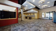 This single-floor space is designed as a well-appointed common area facility, offering a variety of amenities suited for a residential or mixed-use community. The expansive lobby serves as the central hub, featuring multi-toned carpet tiles in earthy grays and reds, structural columns, and a recessed ceiling with layered soffits illuminated by recessed lighting. Bold red accent walls and warm beige tones create a welcoming, modern aesthetic throughout.

A prominent mailroom wall lined with individual metal mailboxes is framed with yellow trim accents, accompanied by dark wood tables for sorting correspondence. Adjacent corridors lead to additional amenities, including a game room furnished with ping pong tables and lounge seating, bathed in natural light from large floor-to-ceiling windows that overlook outdoor greenery. A fully equipped kitchen features oak cabinetry, stainless steel appliances, a full-size refrigerator, and a central island with metal bar stools, alongside convenient restroom access. A dedicated bike storage room provides wall-mounted racks and ample floor space for residents' bicycles. Vending machines and a glazed community room with exterior-facing windows round out the functional, resident-focused layout.