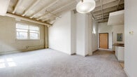 This single-floor loft unit showcases a compelling blend of industrial character and modern finishes throughout. The living area is a standout feature, defined by exposed white-painted brick walls, structural wood beams overhead, and open ceiling elements that lend a raw, warehouse-inspired aesthetic. Neutral carpet flooring runs throughout the main living spaces, softening the industrial tone, while multiple windows allow natural light to filter in and illuminate the textured brick surfaces. A glass-paned exterior door provides additional light and outdoor access from the main living area.

The kitchen is well-appointed with light maple cabinetry offering generous storage, dark laminate countertops, and a full suite of black appliances including a refrigerator, microwave, dishwasher, and range. A peninsula with a built-in cooktop extends the workspace and provides an informal dining or prep area. Tile flooring distinguishes the kitchen from the carpeted living spaces.

A corridor connects the living zone to the private areas of the unit, where the bathroom features warm wood cabinetry, a stone-toned countertop with an undermount sink, a full bathtub with shower, and neutral tile flooring throughout. The overall ambiance balances urban, loft-style architecture with practical, comfortable finishes.