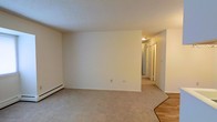 This single-floor space presents a clean, straightforward layout designed for functional living. The main living area features wall-to-wall carpeting in a neutral beige tone, complemented by crisp white walls and a textured ceiling that together create a bright, understated atmosphere. A baseboard heating unit runs along one wall, providing an efficient heating solution integrated seamlessly into the room's perimeter.

From the living area, a hallway extends toward the back of the unit, offering a glimpse of the remaining rooms. The corridor is lined with closet doors, suggesting ample storage options throughout. The hallway is illuminated by a ceiling-mounted light fixture, ensuring the interior remains well-lit. The flooring transitions from carpet in the living space to what appears to be a hard surface near the entryway, adding a practical element at the threshold.

The overall aesthetic is neutral and move-in ready, with a palette of whites and beiges that provides a versatile backdrop for a variety of interior styles. The space conveys a modest, practical character suited to straightforward, comfortable everyday living.