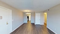 This single-floor space presents a well-maintained and thoughtfully arranged interior designed for comfortable everyday living. The main living area is open and airy, showcasing warm medium-toned hardwood-style flooring that extends seamlessly throughout the space, complemented by crisp white baseboards and soft neutral-toned walls that create a clean, inviting atmosphere. A classic multi-arm chandelier serves as a subtle focal point, adding a touch of traditional elegance to the dining or transitional area. The textured ceiling adds depth and character to the overall aesthetic. A central white door provides access to additional areas of the unit, while an open corridor leads toward what appears to be a kitchen, where black appliances are visible — offering a modern contrast to the light surroundings. A secondary hallway branches off to the right, suggesting additional rooms beyond the main living space. The layout flows naturally, with clear sightlines connecting each area for a cohesive and functional feel. Overall, the space strikes a balance between simplicity and warmth, making it a versatile and welcoming environment suitable for a variety of uses.
