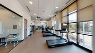 Located at 4111 Broadway Blvd in Kansas City, Missouri, this well-appointed fitness center offers a comprehensive workout environment across a single, expansive floor. The facility is thoughtfully divided into distinct zones to accommodate a wide range of exercise needs.

The cardio area is a standout feature, lined with a generous row of Life Fitness treadmills and stationary cycling bikes positioned directly alongside floor-to-ceiling windows. These large, light-filled windows frame views of the surrounding Broadway Boulevard streetscape, providing an engaging and energizing backdrop during workouts. Roller shades allow for flexible light control throughout the day.

The strength training zone offers a robust selection of cable machines, multi-use functional trainers, free weights, and benches set atop durable rubber flooring. Wall-mounted flat-screen TVs are positioned throughout, enhancing the workout experience. Ceiling fans and recessed lighting throughout the main floor ensure a comfortable and well-lit environment.

A dedicated studio space sets itself apart with warm wood-look flooring, a bold patterned accent wall, and a wall-mounted display screen. This room is stocked with TRX suspension trainers, medicine balls, resistance bands, stability equipment, and a spin bike — ideal for group classes or individual functional training sessions. The overall design is clean, modern, and purposefully organized for an efficient fitness experience.