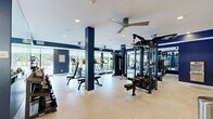 This fitness center offers a generously sized, single-floor workout facility designed to accommodate a comprehensive range of exercise needs. The space is defined by a striking navy blue and white color scheme, with bold accent walls and structural columns that lend a modern, athletic aesthetic throughout.

The cardio zone lines the windowed perimeter, featuring a row of treadmills, an elliptical, a stationary bike, and a stair climber, all positioned to take advantage of natural light and outdoor views of surrounding greenery. Large windows with roller shades allow for adjustable light control while maintaining a bright, open atmosphere.

The strength training area is centrally and laterally arranged, housing multi-station cable machines, a functional training tower with a pull-up bar, and adjustable weight benches. A well-stocked dumbbell rack lines one wall, spanning a wide range of weights to support varied training levels.

Full-length mirrors are strategically placed to visually expand the space and assist with form during workouts. Light wood-tone flooring in a herringbone pattern adds warmth and texture underfoot, while recessed lighting and ceiling fans maintain a comfortable environment. The overall layout is open and uncluttered, allowing for ease of movement between equipment stations.