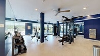This fitness center offers a generously sized, single-floor workout facility designed to accommodate a comprehensive range of exercise needs. The space is defined by a striking navy blue and white color scheme, with bold accent walls and structural columns that lend a modern, athletic aesthetic throughout.

The cardio zone lines the windowed perimeter, featuring a row of treadmills, an elliptical, a stationary bike, and a stair climber, all positioned to take advantage of natural light and outdoor views of surrounding greenery. Large windows with roller shades allow for adjustable light control while maintaining a bright, open atmosphere.

The strength training area is centrally and laterally arranged, housing multi-station cable machines, a functional training tower with a pull-up bar, and adjustable weight benches. A well-stocked dumbbell rack lines one wall, spanning a wide range of weights to support varied training levels.

Full-length mirrors are strategically placed to visually expand the space and assist with form during workouts. Light wood-tone flooring in a herringbone pattern adds warmth and texture underfoot, while recessed lighting and ceiling fans maintain a comfortable environment. The overall layout is open and uncluttered, allowing for ease of movement between equipment stations.