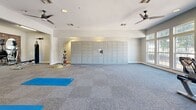 Located at 2702 N Judge Ely Blvd in Abilene, Texas, this well-appointed fitness center spans a generous single floor, offering a comprehensive workout environment designed to accommodate a wide range of exercise needs. The space is divided into distinct functional zones, each thoughtfully equipped and finished.

The main cardio area features a striking exposed brick accent wall with decorative arched mirrors, housing a row of treadmills, a stair climber, and stationary bikes. Wall-mounted televisions enhance the cardio experience, while ceiling fans maintain airflow throughout. A separate strength and functional training zone includes a multi-station cable machine, a weight bench, a punching bag, and a variety of free weights and medicine balls on dedicated racks.

A dedicated stretching and floor exercise area provides ample open space with yoga mats, ceiling fans, and a wall-mounted TV, creating a calm setting for flexibility and low-impact workouts. Large, multi-pane windows along one wall flood this zone with natural light and offer views of the outdoor surroundings.

An additional area houses a full wall of secure package lockers alongside open space suitable for group exercise or additional training. Throughout the facility, neutral-toned carpet tiles, bright flush lighting, and clean white walls create a clean, modern, and motivating atmosphere.