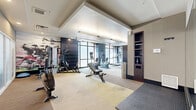 Located at 2401 Chicago Ave in Minneapolis, MN, this modern fitness center offers a thoughtfully designed, single-floor workout facility that provides ample space for a full range of exercise activities. The layout is organized to accommodate multiple workout zones, each serving a distinct fitness purpose.

The cardio area is anchored by a row of treadmills and elliptical machines, all positioned to face large, floor-to-ceiling arched windows that flood the space with natural light and offer open views. Ceiling fans and recessed lighting ensure comfort throughout. Stationary bikes with integrated monitors occupy an additional zone, providing dedicated space for cycling workouts.

The strength training section includes adjustable benches, a multi-station cable and weight machine, and a dedicated free weights area. Built-in shelving units provide convenient storage for accessories and personal items. A bank of lockers along one wall adds practical storage for users.

A separate section with frosted glass partitions and wood-look flooring creates a more private stretching or functional training zone, complete with additional benches and equipment. Motivational fitness imagery is displayed on a feature wall, adding to the energetic atmosphere.

The overall design blends neutral tones, dark accent panels, and modern finishes to create a clean, professional environment well-suited for daily fitness use.