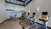 This fitness center is designed to support a comprehensive cardio-focused workout experience. The space is outfitted with a row of treadmills, each equipped with individual monitor screens, positioned alongside elliptical trainers and a stationary bike. The machines are arranged to maximize use of the floor space while maintaining an open, comfortable layout for users.

The room features notably high ceilings with large upper-level windows framed in dark trim, allowing views into an adjacent elevated area and contributing to a sense of openness. Decorative wall sconces with warm-toned shades are mounted along the walls, providing ambient lighting that complements the recessed ceiling fixtures above.

The walls are finished in a neutral light gray, lending the space a clean, modern aesthetic. The flooring consists of a textured, low-pile carpet suited for gym use, offering durability underfoot. The overall design balances functionality with a polished visual presentation, making the fitness center feel both practical and well-appointed for regular use.