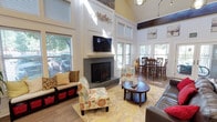 This single-story space is defined by its dramatic vaulted ceilings with warm yellow accents and dark exposed beams, creating an open, airy atmosphere throughout. The main lounge area centers around a stone-accented fireplace flanked by large windows that flood the room with natural light. A wall-mounted television above the fireplace enhances the gathering space, which is furnished with colorful floral accent chairs, a built-in bench with storage, and a round coffee table atop a soft area rug over light hardwood-style flooring.

Adjacent to the lounge, a dining area accommodates both a glass-top round table with dark wood chairs and a separate bar-height dining set, all bathed in natural light from surrounding windows. A dedicated office area features a workstation, desk seating, decorative storage furniture, and a glass-paneled door providing views to an exterior space.

The kitchenette offers a white paneled counter with dark countertop surfaces, pendant lighting, white cabinetry, stainless steel appliances, and a refreshment station. Throughout the space, consistent hardwood-style flooring, white wainscoting, and abundant windows create a cohesive, welcoming environment.