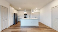 This 1-bedroom, 1-bathroom unit is thoughtfully laid out across a single floor, offering a clean and modern living environment. The open-concept kitchen serves as the focal point of the main living area, featuring white shaker-style cabinetry with gold-toned hardware, bright white countertops, and a full suite of stainless steel appliances including a refrigerator, range, and over-the-range microwave. A large central island with an integrated sink provides additional prep space and casual dining, illuminated by a pair of pendant lights overhead. Wood-look plank flooring flows throughout the main living and kitchen areas, adding warmth to the crisp, neutral palette. The bedroom is a comfortable and well-proportioned space with plush carpet flooring, a ceiling fan, and a window that brings in natural light. It connects directly to a dedicated walk-in closet equipped with built-in shelving and hanging rods for organized storage. The full bathroom features large-format tile flooring, gray cabinetry with gold hardware, a white countertop vanity, and a full soaking tub with shower. Recessed lighting and clean white walls throughout contribute to a bright, airy ambiance that makes the space feel open and inviting.