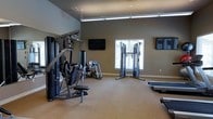 This fitness room offers a functional and organized workout environment finished with neutral gray walls and carpeted flooring. The space is outfitted with a multi-station cable and weight machine, a leg press, and a cable crossover tower, providing a comprehensive range of strength training options. For cardio, at least one treadmill is visible along the perimeter. Two wall-mounted flat-screen televisions are positioned to be visible from various workout stations, enhancing the exercise experience. A window fitted with horizontal blinds allows natural light to enter, contributing to a bright and open atmosphere. Overhead fluorescent lighting ensures consistent illumination throughout. The room is laid out with adequate floor space between equipment, allowing users to move freely and transition between machines with ease. The overall design is practical and straightforward, prioritizing functionality while maintaining a clean, modern aesthetic suited for everyday fitness use.