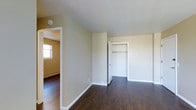This single-floor space features a clean, straightforward layout designed for practical living. The bedroom area is finished with rich, dark hardwood flooring that contrasts neatly against the neutral beige walls and crisp white trim throughout. An open closet fitted with a standard hanging rod provides convenient built-in storage, keeping the room organized and clutter-free. The white-framed closet doors and light ceiling contribute to a bright, airy feel within the space.

A hallway connects to an additional room, visible in the background, which benefits from natural light streaming through a window. The baseboard heating unit lining the wall of the adjoining room is a notable functional feature, ensuring efficient warmth throughout. Vaulted or angled ceiling detailing near the hallway entrance adds subtle architectural interest to an otherwise minimalist interior.

The overall ambiance is clean and understated, with a neutral color palette that provides a versatile backdrop. The consistent use of white trim and doors throughout unifies the space, lending it a cohesive and well-maintained appearance suitable for a variety of furnishing styles.