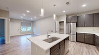 This single-floor unit is designed with a clean, contemporary aesthetic that emphasizes functionality and modern finishes. Light wood-look flooring runs consistently throughout the living areas, complementing the neutral warm-beige wall tones and crisp white baseboards that frame each room.

The kitchen serves as the centerpiece of the open-concept living space, featuring a large center island with a white quartz countertop, an undermount stainless steel sink, and two pendant lights overhead. Dark espresso-toned cabinetry extends along the walls, providing ample storage, and is paired with a full suite of stainless steel appliances including a French door refrigerator, electric range, microwave, and dishwasher. An adjacent open pantry adds additional storage convenience.

The bedrooms are well-proportioned, each featuring windows with roller shades that allow natural light and outdoor views. The primary bathroom offers a dual-sink vanity with backlit mirrors, white quartz countertops, dark cabinetry, and a glass-enclosed walk-in shower.

Additional highlights include a dedicated laundry area with a stacked washer and dryer unit, recessed lighting throughout, and an attached garage with an automatic opener, providing secure and direct access to the unit.