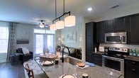 This well-appointed single-floor residence showcases a thoughtfully designed open-concept layout that seamlessly connects the kitchen, dining, and living areas. The kitchen is a standout feature, boasting rich dark cabinetry with ample storage, granite countertops with natural stone veining, a deep undermount sink with a matte black faucet, and stainless steel appliances including a built-in microwave. A striking industrial-style pendant light fixture with three drum shades hangs above the kitchen island, which offers bar seating and serves as a natural divider between the kitchen and living spaces. The living area is warm and inviting, featuring hardwood-style flooring, soft neutral wall tones, and large windows dressed with patterned curtains that allow generous natural light to fill the room. A ceiling fan adds both function and comfort to the space. French doors lead directly to an outdoor balcony, extending the living area and providing an easy indoor-outdoor connection. The overall ambiance is modern yet comfortable, with contemporary finishes and a cohesive neutral palette that creates a bright, airy atmosphere throughout.