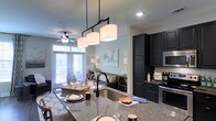 This well-appointed single-floor residence showcases a thoughtfully designed open-concept layout that seamlessly connects the kitchen, dining, and living areas. The kitchen is a standout feature, boasting rich dark cabinetry with ample storage, granite countertops with natural stone veining, a deep undermount sink with a matte black faucet, and stainless steel appliances including a built-in microwave. A striking industrial-style pendant light fixture with three drum shades hangs above the kitchen island, which offers bar seating and serves as a natural divider between the kitchen and living spaces. The living area is warm and inviting, featuring hardwood-style flooring, soft neutral wall tones, and large windows dressed with patterned curtains that allow generous natural light to fill the room. A ceiling fan adds both function and comfort to the space. French doors lead directly to an outdoor balcony, extending the living area and providing an easy indoor-outdoor connection. The overall ambiance is modern yet comfortable, with contemporary finishes and a cohesive neutral palette that creates a bright, airy atmosphere throughout.