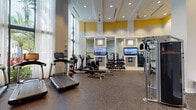 This fitness center is designed to support a comprehensive workout experience within a thoughtfully appointed, single-floor facility. Soaring ceilings and floor-to-ceiling windows along one wall flood the space with natural light, while sheer white curtains soften the brightness and offer views of lush greenery outside. Bold yellow accent panels near the upper walls add a vibrant, energetic touch to the otherwise clean white and gray interior palette.

The main workout floor is finished with durable rubber flooring throughout, providing both safety and comfort underfoot. Cardio equipment is well-represented, with a row of treadmills positioned along the windowed wall, complemented by elliptical trainers and stationary cycling bikes. Multiple flat-screen televisions are mounted at eye level to keep users entertained during workouts.

The strength training area features a tiered dumbbell rack stocked with a full range of weights, a barbell and plate tree for free weight exercises, a multi-function cable machine, and built-in shelving for additional storage. Mirrored wall panels expand the sense of space and allow users to monitor their form.

Near the entrance, a vending machine offers convenient refreshment access. The facility is accessible through double glass-paneled doors and benefits from an open, airy layout that encourages movement and ease of use throughout.
