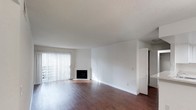 This single-floor residence offers a clean and functional layout with a bright, open living area at its heart. The living room is finished with rich, dark hardwood flooring that contrasts beautifully against crisp white walls, creating a timeless and inviting atmosphere. A corner fireplace serves as a subtle focal point, adding warmth and character to the space. Floor-to-ceiling vertical blinds frame a set of sliding glass doors, allowing an abundance of natural light to flood the interior while providing access to an outdoor area. The smooth, neutral-toned ceiling complements the overall airy feel of the room. Adjacent to the living area, louvered closet doors hint at well-designed storage solutions, while a partially visible adjoining room suggests a practical and connected floor plan. The overall ambiance is calm and understated, offering a versatile canvas well-suited to a variety of interior styles. The space balances openness with defined zones, making it both comfortable and easy to personalize.