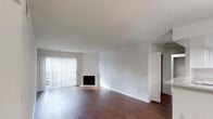 This single-floor residence offers a clean and functional layout with a bright, open living area at its heart. The living room is finished with rich, dark hardwood flooring that contrasts beautifully against crisp white walls, creating a timeless and inviting atmosphere. A corner fireplace serves as a subtle focal point, adding warmth and character to the space. Floor-to-ceiling vertical blinds frame a set of sliding glass doors, allowing an abundance of natural light to flood the interior while providing access to an outdoor area. The smooth, neutral-toned ceiling complements the overall airy feel of the room. Adjacent to the living area, louvered closet doors hint at well-designed storage solutions, while a partially visible adjoining room suggests a practical and connected floor plan. The overall ambiance is calm and understated, offering a versatile canvas well-suited to a variety of interior styles. The space balances openness with defined zones, making it both comfortable and easy to personalize.