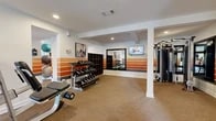 This fitness center offers a generously sized, single-floor layout thoughtfully designed to accommodate a full range of workout activities. The exercise room is the centerpiece of the space, featuring a striking interior design highlighted by bold horizontal orange stripe accents along the white walls, creating an energetic and motivating atmosphere. Recessed lighting throughout ensures consistent, bright illumination, while large windows along one wall flood the space with natural light and provide views of the surrounding greenery.

The exercise room is well-appointed with a diverse selection of equipment, including multiple treadmills, an elliptical machine, a multi-station cable machine, a dedicated free weights area with a tiered dumbbell rack, an adjustable weight bench, and stability balls. Wall-mounted flat-screen televisions are positioned for visibility from both the cardio and strength training zones. Large framed mirrors enhance the sense of openness and allow users to monitor their form during workouts. Structural columns are cleanly integrated into the layout without impeding the generous open floor area, which is covered in durable, neutral-toned carpet.

The facility also includes a dedicated bathroom finished with classic white subway tile wainscoting, large-format floor tiles, bold orange upper walls, and accessibility-compliant fixtures including grab bars and ample maneuvering space.