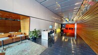 Located in Portland's vibrant urban core, this polished commercial lobby presents a seamless blend of contemporary design and warm natural materials. The entrance corridor is defined by large-format slate tile flooring with a high-gloss finish that reflects the ambient lighting above, creating a sense of depth and sophistication throughout the space. Soaring ceilings with reflective paneling amplify the openness of the floor plan, while a glass clerestory draws in natural light from the street-facing facade.

At the heart of the lobby stands a striking reception desk crafted from rich warm wood with illuminated base detailing, anchoring the space and providing a welcoming focal point. Adjacent to the reception area, a collaborative workstation with a wall-mounted display and ergonomic bar-height seating offers a functional area for meetings or check-ins. A glass-enclosed lounge nook, finished in natural wood slat paneling and furnished with leather banquette seating and bistro tables, provides a comfortable retreat.

Further into the space, bold artisan wall art with geometric, colorful patterns crowns a full-length horizontal wood slat feature wall, adding artistic character. A dedicated package locker bank offers practical amenity storage. Frosted glass partitions and warm accent lighting define ancillary service corridors, maintaining privacy while contributing to the overall refined ambiance of this thoughtfully designed lobby.