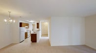 This single-story space features an open-concept design that seamlessly connects the kitchen and living area, creating a cohesive and functional floor plan. The kitchen is equipped with warm wood-toned cabinetry, white appliances including a refrigerator and electric range, and a built-in peninsula that provides additional counter space and serves as a natural divider between the kitchen and the adjoining living area. A overhead fluorescent light fixture illuminates the kitchen workspace efficiently. The living area is finished with neutral-toned carpeting that extends throughout, contributing to a clean and consistent aesthetic. White walls throughout the space provide a bright, airy feel and offer versatility for personalization. A hallway visible beyond the kitchen area leads to additional rooms, suggesting a practical layout designed for easy navigation. The overall ambiance is understated and functional, with a straightforward design that prioritizes usability. The open sightlines between the kitchen and living space enhance the sense of roominess, making the unit feel welcoming and easy to move through.