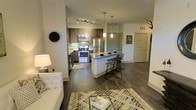 This single-floor space showcases a thoughtfully designed open-concept layout that integrates the kitchen, dining, and living areas into one cohesive, functional environment. The kitchen is outfitted with stainless steel appliances, including a gas range, microwave, and refrigerator, complemented by warm wood-toned cabinetry and a decorative tile backsplash. Track lighting illuminates the cooking area, while pendant lights hang above the central island, which features a granite countertop and accommodates bar-style seating with modern metal stools — making it ideal for casual dining or entertaining. The island serves as a natural dividing point between the kitchen and the open living area, which is anchored by rich, dark hardwood-style flooring that runs throughout the space. The living area offers ample room for comfortable furnishings and benefits from a bright, neutral color palette that enhances the sense of openness. Additional doorways visible in the background suggest further private rooms beyond the main living space. Overall, the design balances contemporary aesthetics with practical functionality.
