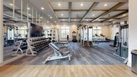 Located at 401 Nonaville Rd in Mount Juliet, Tennessee, this well-appointed fitness center is designed to support a comprehensive workout experience in a bright, welcoming environment. The space is organized across a single floor and divided into distinct functional zones, allowing for both strength training and cardiovascular exercise.

The interior is defined by its striking coffered ceilings adorned with rustic wood beam accents, recessed lighting, and ceiling fans that contribute to a comfortable atmosphere during workouts. Large windows line multiple walls, inviting generous natural light and offering views of the surrounding outdoor scenery.

The strength training area is thoroughly equipped with a full rack of dumbbells, adjustable benches, cable machines, a multi-functional cable crossover system, and additional selectorized strength machines. A wall-mounted flat-screen TV and full-length mirrors enhance the experience in this zone. A motivational mural reading "Let's Get That Sweat On!" adds an energetic touch to the space.

The cardio zone features a row of treadmills, stair climbers, elliptical machines, and stationary bikes, all positioned near the windows to maximize natural light during workouts. The flooring throughout combines durable, textured carpet tiles in the equipment areas with smooth hardwood-style flooring in transitional zones, ensuring both functionality and aesthetic cohesion throughout the facility.