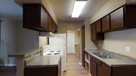 This two-floor residence offers a practical and well-organized layout designed for comfortable living. The kitchen is a standout feature, styled in a classic galley configuration that maximizes efficiency within the space. Dark espresso-toned cabinetry lines both sides of the kitchen, providing generous storage both above and below the countertops. The countertops display a granite-style finish, adding a touch of texture and visual interest to the space. Essential appliances are included, among them a white electric coil-top range and a full-size refrigerator, making the kitchen move-in ready. A stainless steel sink is positioned along one wall, complemented by additional open shelving for easy-access storage. The kitchen is illuminated by a central overhead fluorescent light fixture that brightens the entire cooking area. Warm-toned wood-style flooring flows through the kitchen and into the adjacent areas, lending continuity and a welcoming feel throughout the home. An arched passageway at the end of the kitchen leads further into the residence, hinting at additional living and utility spaces beyond. The overall ambiance of the home is practical and homey, with a neutral color palette that provides a versatile backdrop for personalization across both floors.