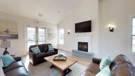 This single-story community room is thoughtfully designed to serve as a versatile gathering space, combining comfort and functionality throughout its well-appointed areas. The interior is unified by light wood-look flooring, crisp white walls, and vaulted ceilings that lend an open, airy quality to the entire space.

The welcoming entry foyer features soaring ceilings and a decorative console table with a large framed mirror, setting a warm, coastal-inspired tone. The main lounge area centers around a classic white-mantled fireplace with a wall-mounted flat-screen television above, complemented by rich brown leather seating arranged around a rustic wooden coffee table. Coastal artwork and wall sconces add character throughout.

An adjacent recreation area accommodates a full-size billiards table beneath generous vaulted ceilings, with additional high-top seating nearby for casual socializing. The kitchenette area offers a stainless steel refrigerator and a high-top bistro table with bar stools beside a large window with outdoor views.

Multiple sets of glass double doors provide direct access to an outdoor patio area, allowing natural light to flow freely into the space. Large windows throughout further enhance the bright, welcoming ambiance of this well-equipped community room.