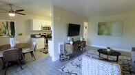 This single-floor residence offers a thoughtfully designed open-concept layout that seamlessly connects the living area and kitchen, creating a spacious and functional environment. The living room is bright and welcoming, featuring vaulted ceilings that enhance the sense of openness. Large sliding glass doors flood the space with natural light and provide direct access to a private balcony overlooking mature trees and greenery, bringing a serene outdoor connection to the interior. The living area is styled with warm, neutral tones complemented by rich accent decor, including a bold oversized art piece that adds a sophisticated focal point to the room. Hardwood-style flooring runs throughout, lending a clean and cohesive look to the space. The kitchen is positioned adjacent to the living area and features white cabinetry, dark countertops, and stainless steel appliances, offering both style and practicality. A half-wall partition subtly defines the kitchen from the main living space while maintaining an open, airy feel. A wall-mounted flat-screen television and media console anchor the living room's entertainment zone. Overall, the space presents a modern, comfortable ambiance ideal for both relaxed living and everyday functionality.