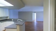 This single-floor space showcases an open-concept design that seamlessly connects the kitchen and living areas, creating a sense of spaciousness and easy flow. The kitchen features a peninsula with a dark granite-style countertop and an integrated sink, providing both functional workspace and a natural visual divider between the cooking and living zones. White cabinetry beneath the peninsula adds a clean contrast to the darker countertop surface.

The flooring throughout consists of dark hardwood-style planks that lend warmth and continuity across the open layout. Neutral gray walls create a calm, versatile backdrop suitable for a variety of interior styles. Recessed lighting is installed in the ceiling, supplementing the natural light that appears to enter from an adjacent room or hallway visible in the background.

The open hallway leading off the main living area suggests additional rooms beyond, offering private spaces separate from the communal living zone. A ceiling-mounted HVAC vent ensures climate control throughout. Overall, the space presents a clean, minimalist aesthetic with practical finishes, making it a functional and adaptable living environment.