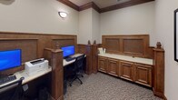 This single-floor business center is thoughtfully designed to support productivity in a refined, professional setting. The space is distinguished by its rich warm-toned wood millwork throughout, including raised-panel wainscoting, decorative pilasters with finial caps, and built-in cabinetry that lend the environment a polished, classic character.

Workstations are seamlessly integrated into the design, featuring dedicated computer terminals with monitors, keyboards, and ergonomic seating, each positioned within semi-private wood-paneled alcoves that encourage focused work. A built-in credenza with paneled cabinet doors provides additional surface space and storage, keeping the area organized and functional.

The flooring transitions from a patterned carpet in the main work area to stone tile in the connecting corridor, adding visual texture and clear spatial definition. Neutral wall tones complement the woodwork and create a calm, professional atmosphere.

Natural light filters in through windows visible from the hallway entrance, while wall-mounted sconces provide warm supplemental lighting within the work area. Framed artwork adorns the walls, adding a touch of sophistication. The layout allows for ease of movement between the workstations and the entrance, making this business center an efficient and elegantly appointed space for guests or professionals needing a dedicated workspace.