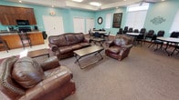 This single-floor community space is designed to accommodate both relaxed gatherings and organized events. The interior is defined by its signature aqua-toned walls, which create a bright, welcoming atmosphere throughout. The main lounge area is generously proportioned, featuring tray ceilings with ceiling fans and flush-mounted lighting, plush brown leather sofas and armchairs arranged around folding coffee tables, and carpeted flooring complemented by decorative area rugs. A striking stone fireplace with a wood mantle serves as a focal point, flanked by tall tropical plants and framed artwork that enhance the room's character. Multiple round tables with stackable chairs are positioned throughout the space, accommodating various group configurations for meetings or events. A wall-mounted television adds functional utility to the lounge. The fully equipped kitchen features warm wood cabinetry, black appliances including a refrigerator, range, and dishwasher, a large central island with bar seating, granite-style countertops, and light tile flooring. Windows fitted with blinds allow natural light to filter throughout, contributing to the overall open and airy feel of the space.