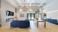 The Trailside Apartments clubhouse in Boylston, MA is a thoughtfully designed community amenity space spread across a single expansive floor. The welcoming entrance foyer features wide-plank light hardwood floors, soaring coffered ceilings, and geometric pendant chandeliers that set an inviting tone throughout. The main lounge area is anchored by a bold navy sectional sofa, a modern linear fireplace, and a wall-mounted television, complemented by large glass doors that frame wooded views and flood the space with natural light. Adjacent to the lounge is a communal kitchen and dining area, outfitted with navy cabinetry, built-in appliances, open shelving with under-cabinet lighting, and a substantial wood communal table — ideal for resident gatherings. A flex lounge nook with vibrant patterned carpeting, curated artwork, and a bistro-style table provides a quieter setting for small meetings or casual conversation. A games room with a shuffleboard table and accent seating adds a recreational dimension. The fitness center is generously proportioned, featuring a full array of cardio machines, free weights, stability equipment, and a dedicated yoga and stretching studio — all finished with eye-catching herringbone-patterned flooring and large windows overlooking the surrounding greenery.