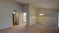 This single-story residence showcases a spacious, open-concept layout defined by striking vaulted ceilings that lend an airy, expansive feel to the main living area. The generous ceiling height draws the eye upward and enhances the sense of volume throughout the space. Neutral gray walls and wall-to-wall carpet in a warm beige tone provide a clean, understated backdrop adaptable to a variety of interior styles.

A decorative chandelier hangs prominently over the dining area, adding a touch of elegance to the otherwise understated design. Adjacent to the dining space, a built-in pass-through with a countertop niche connects seamlessly to the kitchen, facilitating ease of entertaining and daily use.

The kitchen, visible through an open doorway, features white cabinetry, a sink, and under-cabinet lighting, offering a bright and functional workspace. A hallway off the main living area leads to additional rooms, with a bi-fold closet door visible, suggesting convenient storage options. A wall-mounted thermostat indicates climate control throughout. Overall, the space presents a well-proportioned, move-in-ready layout with cohesive finishes.