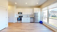 This clean, modern single-floor space is thoughtfully designed with an open-concept layout and a cohesive aesthetic throughout. Light wood-tone flooring flows seamlessly from room to room, creating a warm and unified feel across the entire unit.

The kitchen is a standout feature, showcasing white shaker-style cabinetry that extends to the ceiling for generous storage, complemented by a white subway tile backsplash and light stone countertops. A full suite of stainless steel appliances includes an electric range, microwave, dishwasher, and refrigerator. Large windows adjacent to the kitchen flood the space with natural light and offer outdoor views, enhancing the open and airy atmosphere.

The living area is spacious and bright, with recessed lighting and a window that further contributes to the light-filled environment. The neutral warm-white walls provide a versatile backdrop suitable for a variety of interior styles.

The bathroom is well-appointed with a floating white vanity featuring ample drawer storage, a white countertop with an integrated sink, a full tub-shower combination, and chrome fixtures throughout. The consistent wood-tone flooring continues here, maintaining the unit's cohesive design language.

A hallway with built-in storage panels and recessed lighting connects the rooms efficiently, rounding out a well-organized and functional floor plan.