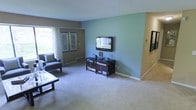 This single-story space showcases a well-appointed living area designed with comfort and functionality in mind. The room features a soothing two-tone color scheme, pairing a soft teal accent wall with warm taupe walls, creating a balanced and inviting ambiance. Plush, light-toned wall-to-wall carpeting runs throughout, adding warmth underfoot and contributing to the room's relaxed atmosphere.

A large window along one wall allows natural light to filter in, brightening the space and offering a view of the surrounding greenery. Sheer white curtains frame the window, softening the light while maintaining an open feel. Two framed botanical prints are displayed nearby, adding a decorative touch.

The living area is anchored by a wall-mounted flat-screen television positioned on the teal accent wall, paired with a dark wood media console below that provides ample storage through its shelves and drawers. A glass-topped coffee table and a cushioned armchair complete the seating arrangement, offering a functional and comfortable layout. Subtle architectural details, including clean white baseboards and a smooth ceiling, give the space a tidy, well-maintained character throughout.