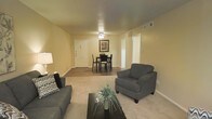 This single-floor residence offers a comfortable and functional living environment designed with everyday convenience in mind. The open-concept living and dining area serves as the heart of the home, seamlessly connecting the two spaces for easy interaction and flow. Warm beige walls and a neutral color palette create an inviting atmosphere throughout, complemented by plush wall-to-wall carpeting in the living area that transitions to smooth hard flooring in the dining space. The living room provides ample space for seating, as evidenced by the generously sized sofa and armchair arrangement centered around a sleek coffee table. A ceiling light fixture illuminates the space with a soft, ambient glow. The adjacent dining area accommodates a compact table and chair set, ideal for everyday meals, and is accented by a framed piece of wall art that adds a touch of personality to the space. A white entry door with visible hardware provides a clean, classic focal point, while multiple HVAC vents integrated into the walls and ceiling speak to the practical infrastructure of the home. Hallway access visible from the main living area suggests additional rooms beyond, completing this thoughtfully arranged and move-in-ready space.
