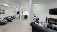 This single-floor space offers an open-concept layout that combines a living area, kitchen, and dining zone into one cohesive and functional environment. The interior is finished with crisp white walls and light wood-style flooring, creating a bright and airy atmosphere throughout. Structural columns punctuate the space, adding architectural character while subtly defining each zone without the use of dividing walls.

The living area is anchored by a tufted dark charcoal sofa accented with decorative pillows, complemented by a patterned accent chair nearby. Recessed lighting and track lighting illuminate the space evenly, enhancing its clean, modern feel. A flat-screen television is mounted in the corner near white cabinetry, which provides ample storage and contributes to the polished look of the space.

The kitchen features a central island with bar-height seating, pendant lighting overhead, and white cabinetry that aligns with the overall neutral palette. Adjacent to the kitchen, a dining area with bar-style seating offers additional space for gathering. The open flow between all areas makes this space well-suited for both everyday use and entertaining, with a consistent modern, neutral design theme carried throughout.