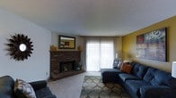 This single-floor residence offers a comfortable and well-appointed living space designed with both functionality and warmth in mind. The living room serves as the heart of the home, anchored by a classic corner brick fireplace complete with a traditional wood mantel — a standout architectural feature that adds character and charm to the room. Above the fireplace, a mirror reflects light and adds depth to the space. Large sliding glass doors flood the room with natural light, framed by patterned sheer curtains that soften the brightness while maintaining an airy feel. An accent wall in a bold mustard yellow adds a pop of color and visual interest, complementing the neutral tones found throughout the rest of the space. The living area is carpeted, contributing to the room's cozy ambiance, and is generously sized to accommodate a large sectional sofa and area rug. The textured ceiling adds a subtle design element consistent with the home's classic style. Overall, the space presents a warm, inviting atmosphere well-suited for everyday living and relaxed entertaining.
