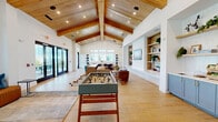 Located at 640 Lakeside Drive in Canton, GA, this expansive single-floor community space offers an impressive blend of modern design and warm, inviting character. The centerpiece of the space is a dramatic vaulted ceiling adorned with exposed natural wood beams and tongue-and-groove paneling, lending a sense of grandeur and warmth throughout. Light hardwood-style flooring runs seamlessly across the entire floor, enhancing the open, airy feel.

The living room is generously proportioned, featuring multiple seating arrangements with leather and upholstered furnishings, built-in shelving niches with a mounted television, and a foosball table for recreation. Large black-framed glass doors open to an outdoor patio, flooding the space with natural light and offering views of the surrounding community. A striking entry foyer showcases an ornate stained-glass front door beneath the soaring ceiling peak.

The dining area offers a vibrant contrast, highlighted by teal-painted cabinetry, iridescent stacked tile, and a wall-mounted display. Built-in banquette seating with blush upholstery, gold-accented wall sconces, and individual café-style tables create an intimate yet functional setting. Curated artwork and thoughtful décor throughout tie the design together into a cohesive, upscale communal environment.