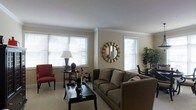 This single-floor space offers a warm and inviting atmosphere highlighted by a well-appointed living area. The room is finished with smooth white ceilings accented by clean crown molding, lending a classic and polished character to the space. Neutral warm-toned walls create a versatile backdrop that complements the carefully chosen furnishings, including a plush taupe sofa, a bold red accent chair, and a sleek dark-finished coffee table that anchors the seating arrangement. A decorative sunburst mirror serves as a focal point on the wall, adding a stylish and refined touch to the overall aesthetic. Multiple windows fitted with white horizontal blinds allow generous natural light to fill the room, creating a bright and airy ambiance throughout the day. Lush indoor greenery in the corner introduces a refreshing organic element, while a classic table lamp and carefully placed decorative accents contribute to the space's layered, curated feel. The flooring is light-toned, enhancing the sense of openness and cleanliness. Overall, this space strikes a thoughtful balance between comfort and style, making it an ideal setting for both relaxed everyday living and entertaining guests.