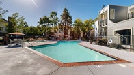 Located at 2850 Middlefield Rd in Palo Alto, California, this outdoor amenity area serves as a central gathering space within a well-maintained residential community. The centerpiece of the space is a generously sized swimming pool with clear, blue-green water, framed by brick-edged coping and a broad concrete sun deck. Adjacent to the pool, a dedicated amenity building features white lap siding, multiple windows, and secured entry doors, providing residents with convenient access to supporting facilities. The surrounding deck extends into a spacious patio area furnished with multiple seating arrangements — including chairs, tables, and vibrant orange market umbrellas — ideal for outdoor dining and relaxation. A separate spa or wading area is also visible near the main pool. The space is enclosed by a black wrought-iron fence with gated entry points, ensuring privacy and security. Mature trees line the perimeter, providing natural shade and a lush backdrop against the California sky. The multi-story residential buildings surrounding the courtyard feature gray horizontal lap siding, covered balconies, and landscaped garden beds with colorful plantings, contributing to the cohesive and welcoming character of the community's shared outdoor environment.
