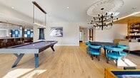 Located at 22 Via Toscana in Salem, New Hampshire, this expansive amenity floor spans a generously proportioned single level, offering a sophisticated blend of social and recreational spaces finished to an exceptional standard throughout.

The interiors are unified by wide-plank light hardwood flooring, recessed lighting, and crisp white walls that lend an airy, contemporary feel. A grand lounge area serves as a central gathering point, anchored by a dramatic marble fireplace surround with a linear gas fireplace, a flatscreen display above, and cove-lit tray ceilings. Floor-to-ceiling windows and sliding glass doors flood the space with natural light while framing panoramic views of the surrounding cityscape.

A dedicated games room features a billiards table, a shuffleboard table, and a round game table with teal upholstered seating beneath a striking multi-arm chandelier with an ornate circular ceiling medallion. A well-stocked built-in library wall with warm shelving adds a refined lounge ambiance to this area.

A sleek bar area showcases a richly textured accent wall, bar seating, pendant lighting, and a mounted display, ideal for entertaining. The hallway entrance is distinguished by a warm wood-paneled ceiling, curated artwork, and plush modern seating areas.

Two balconies provide outdoor access with metal railing and open views over the vibrant downtown streetscape below.