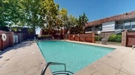 Located at 4201 Meadowlark Ln SE in Rio Rancho, New Mexico, this outdoor pool area serves as a central community amenity set against the backdrop of a classic brick residential building. The generously sized rectangular swimming pool features crystal-clear turquoise water, with entry points provided by stainless steel handrails and ladders on multiple sides for convenient access.

The pool deck is finished in light concrete and offers ample space for relaxation, lined with blue lounge chairs and chaise lounges ideal for sunbathing beneath New Mexico's abundant sunshine. A shaded seating area with an umbrella table provides a comfortable retreat from the sun on warmer days.

The area is enclosed by a combination of iron fencing and brick wall sections, ensuring both security and privacy while maintaining an open, welcoming atmosphere. A safety ring buoy is mounted prominently within the enclosure. Mature deciduous and evergreen trees surround and shade portions of the deck, adding natural beauty and a cooling canopy overhead.

The adjacent multi-story brick building features balconies and staircase access, complementing the warm, earthy architectural character of the complex. The overall ambiance is inviting and resort-like, offering residents a well-maintained outdoor escape well-suited to the sunny climate of the Rio Rancho region.