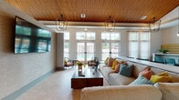 Located in Bradenton, Florida, this single-story community clubhouse at 1161 3rd Ave E is a well-appointed gathering space designed with a coastal-contemporary aesthetic. The interior is anchored by striking vaulted wood-plank ceilings that lend warmth and architectural character throughout, complemented by geometric lantern-style pendant lighting that adds an elegant, modern touch.

The main lounge area is expansive and inviting, furnished with plush neutral sofas accented by vibrant throw pillows, sleek accent chairs in soft blue tones, and a bold yellow ottoman that injects a playful pop of color. A large wall-mounted flat-screen television serves as a focal point, set against a textured stone accent wall. Floor-to-ceiling French doors and transom windows flood the space with natural light while offering direct views of the outdoor pool area.

The kitchen area is thoughtfully designed with white cabinetry, stainless steel appliances including a French door refrigerator, rich hardwood flooring, and a striking stacked stone backsplash wall adorned with sculptural metallic fish art — a nod to the coastal surroundings. A central island with seating provides a casual dining and social hub. Throughout the space, large windows maintain a bright, airy atmosphere, making this clubhouse an ideal venue for community gatherings and relaxation.